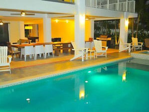 Un espace piscine extérieur moderne, comprenant une table et des chaises pour les repas, un coin bar et un salon au bord de la piscine avec des chaises blanches.