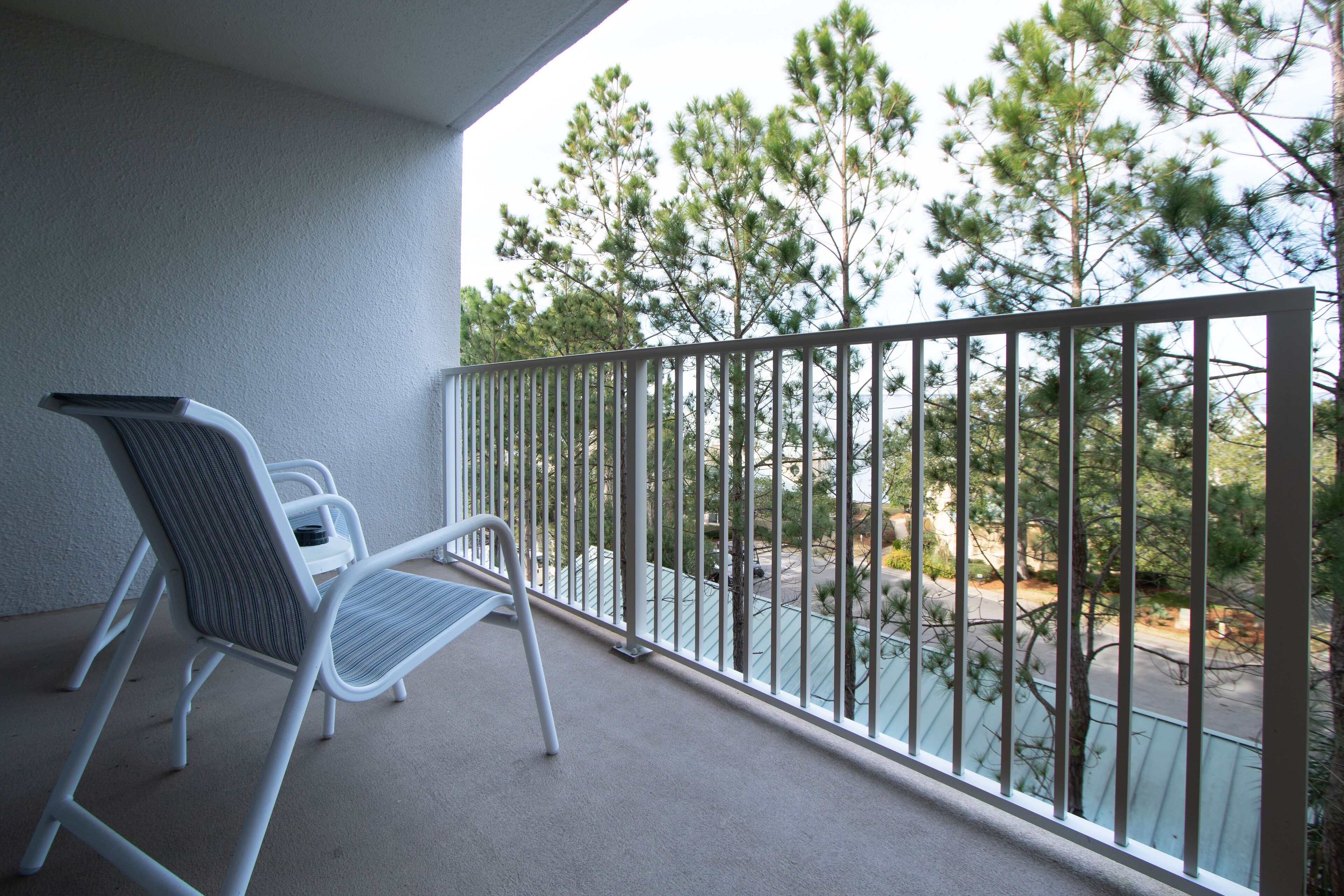 Un balcon avec une balustrade en métal, deux chaises blanches et une vue sur des arbres et une route.