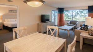 Habitación de hotel con cama, televisor en mueble bajo, mesa de comedor con sillas y vista a árboles por la ventana.