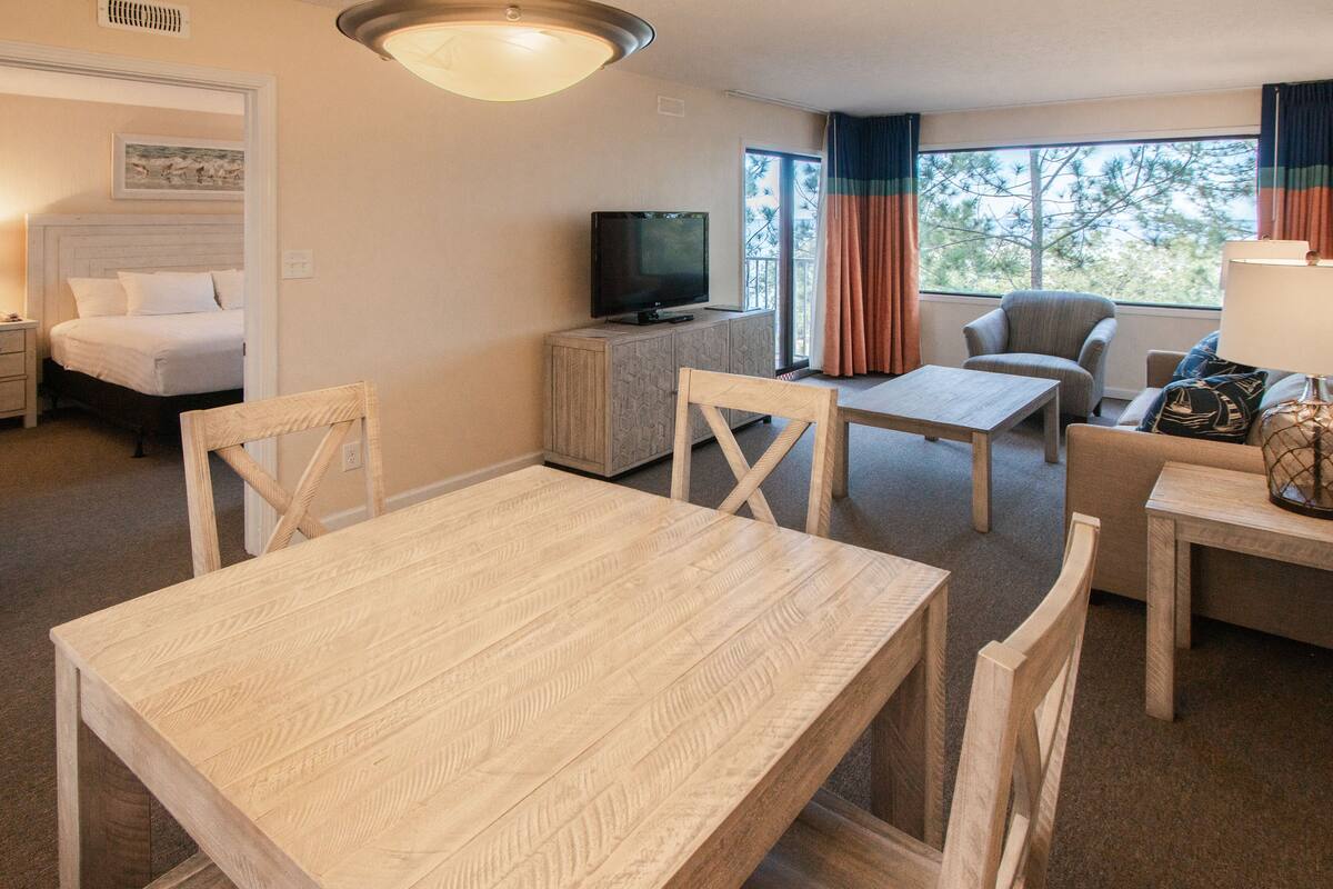 Une chambre d’hôtel avec un lit, un téléviseur posé sur un meuble, une table à manger avec des chaises, et une vue sur les arbres par la fenêtre.