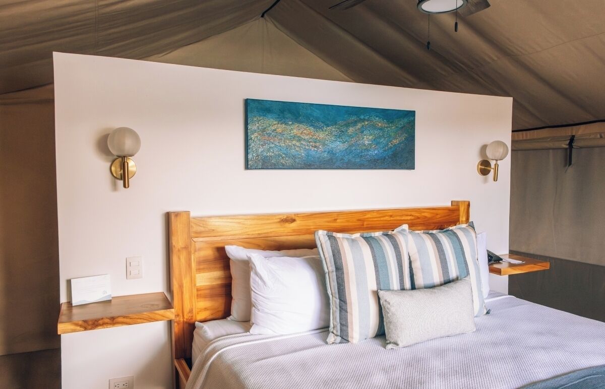 A bedroom with a wooden headboard, a bed with striped bedding, two wall-mounted lamps, and a blue abstract painting.
