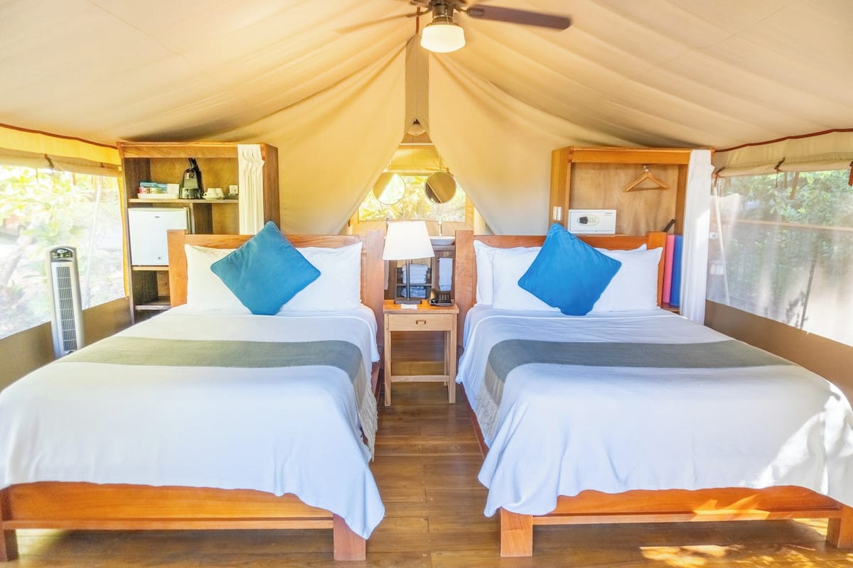 A tented accommodation with two beds, a small table with a lamp, and a kitchenette.