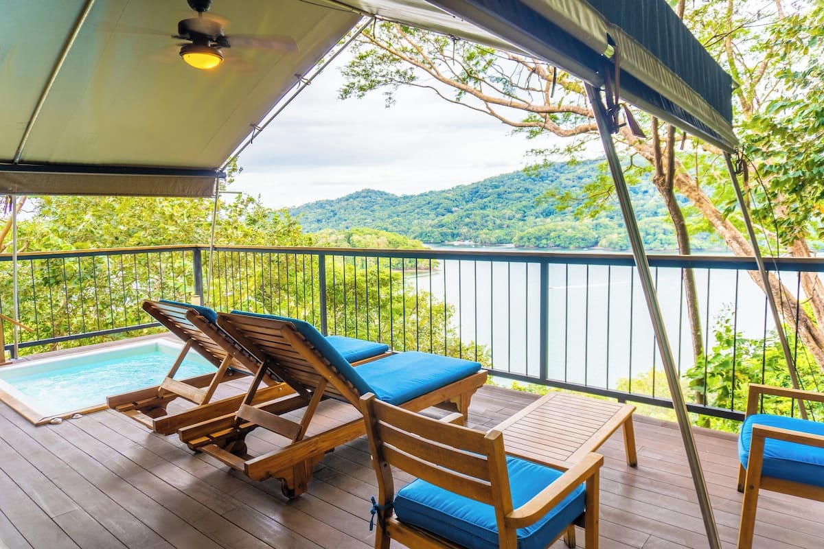 A wooden deck with lounge chairs and a table, overlooking a body of water and green hills.