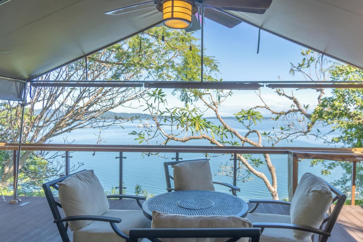 A covered outdoor seating area with a table and chairs, overlooking a body of water and trees.