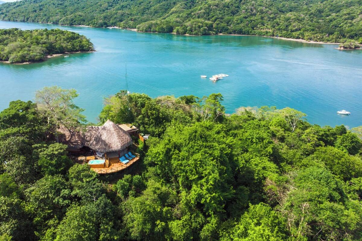 A tropical resort with a thatched-roof hut and a wooden deck with blue lounge chairs, surrounded by lush greenery and a turquoise lake.