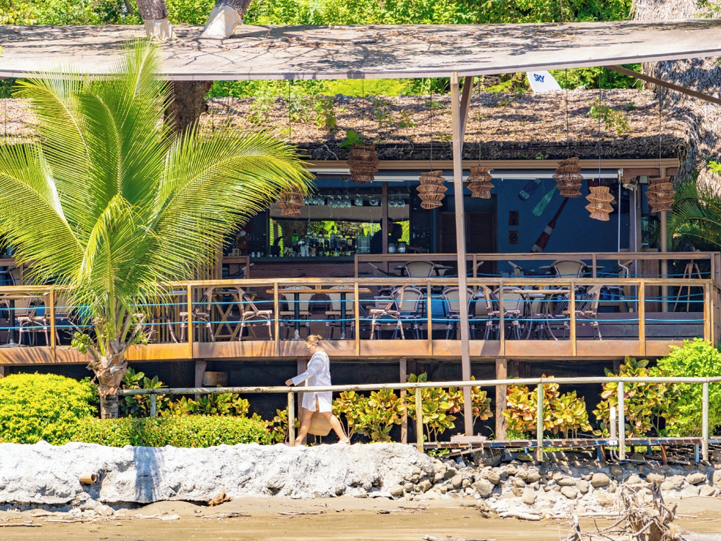 Un restaurante en la playa con terraza, una palmera y una persona caminando.