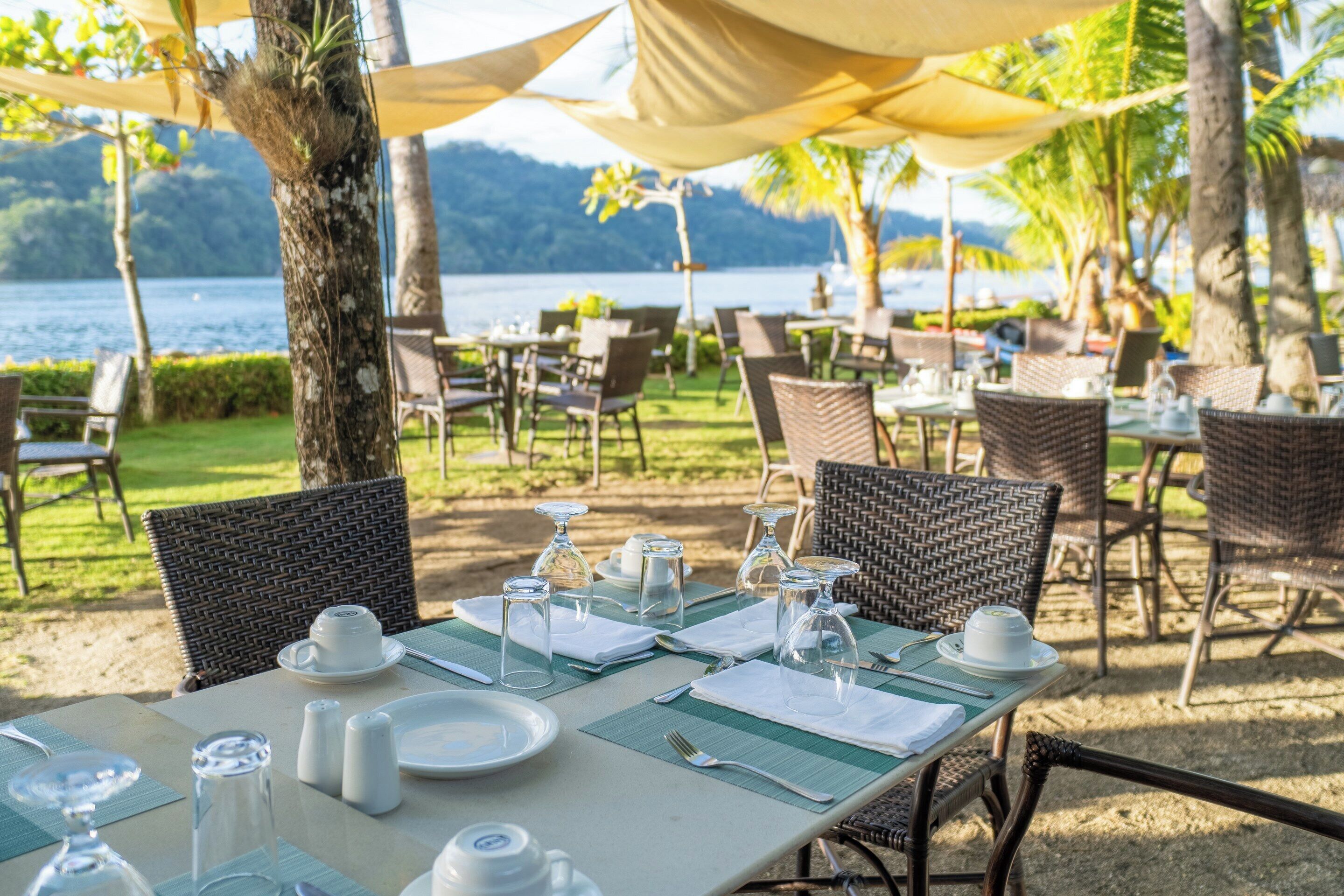 An outdoor dining area with tables set for two, surrounded by wicker chairs, with a view of a lake and mountains in the background.
