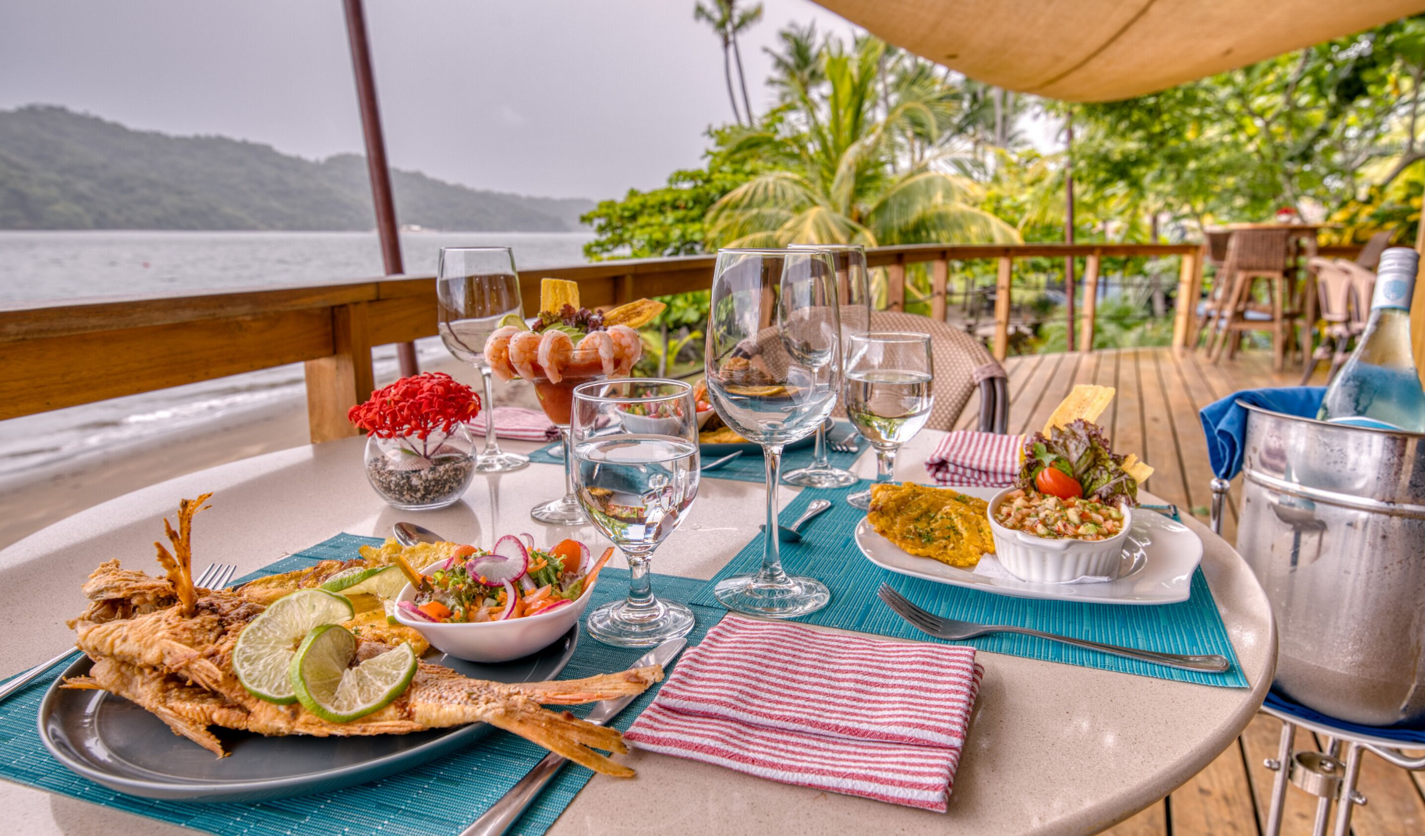 A table set with seafood, drinks, and tropical decor on a deck overlooking the sea.