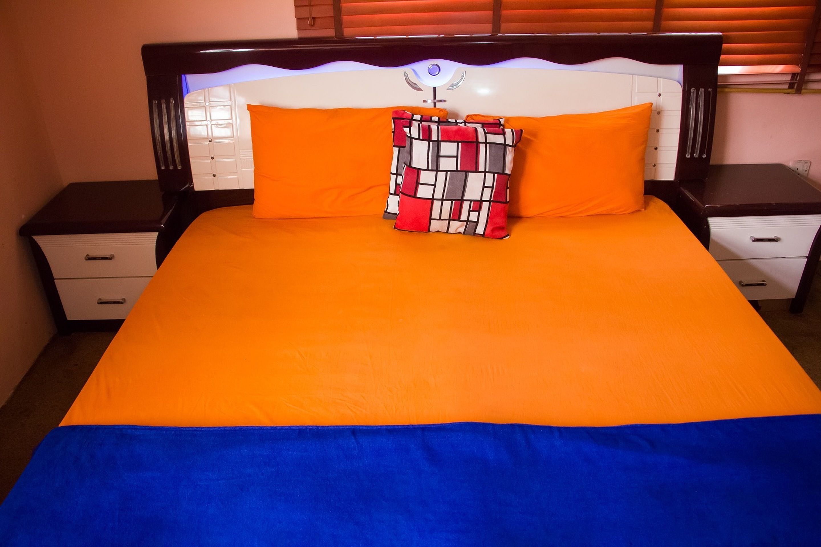A bed with an orange and blue bedspread, a patterned pillow, and a headboard with a light fixture.