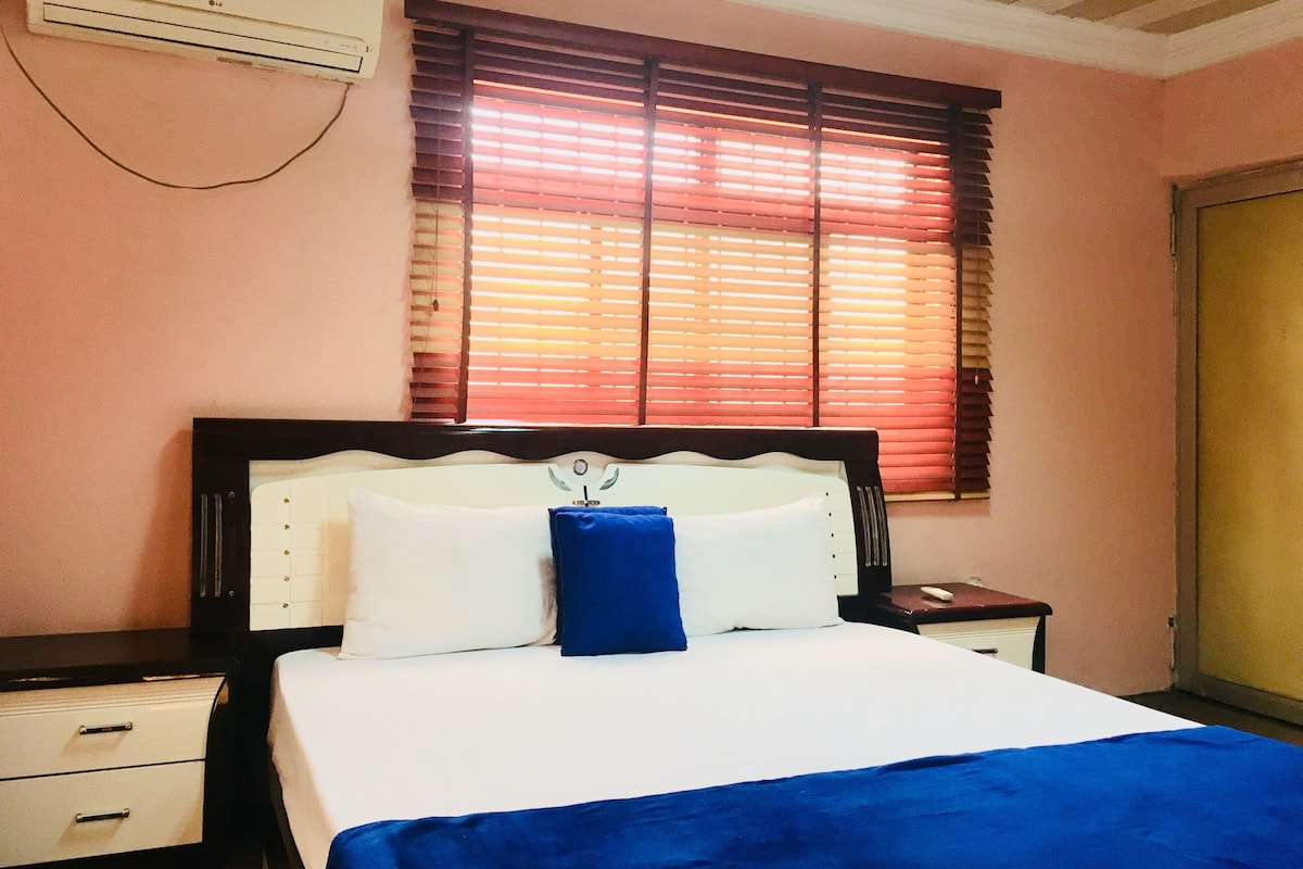 A hotel room with a bed, a nightstand, a drawer unit, and a window with blinds.