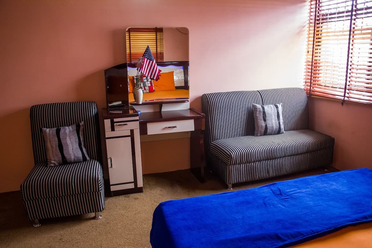 A hotel room with a bed, two striped chairs, a desk with a mirror, and a window with blinds.
