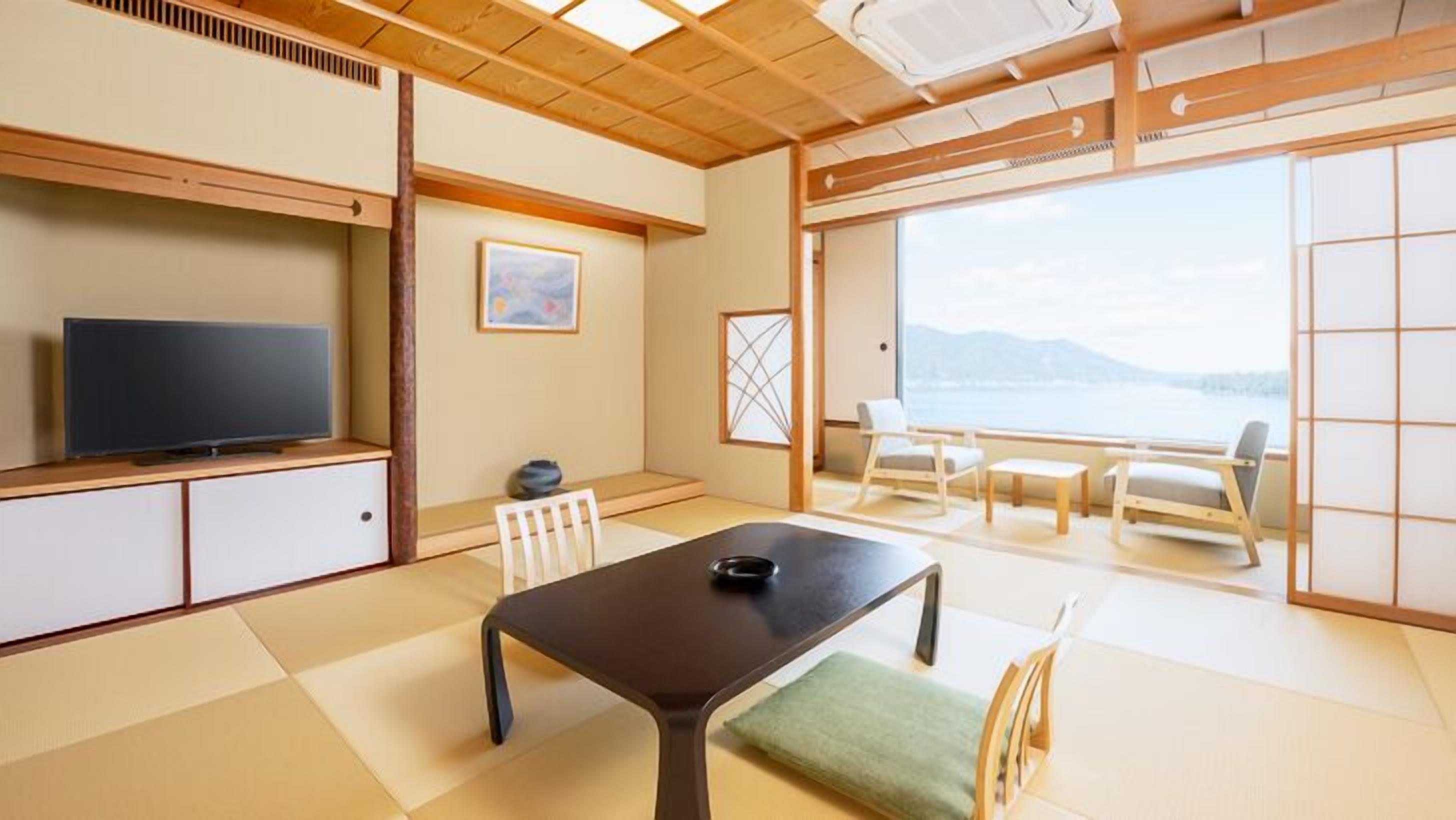 A traditional Japanese-style room with tatami flooring, a television, a table with a green cushion, and a view of the sea through large windows.