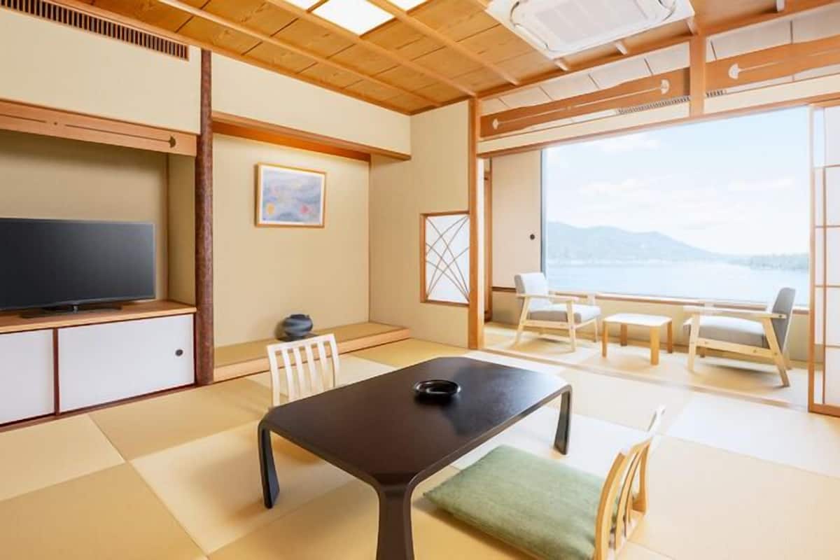 A traditional Japanese-style room with tatami flooring, a television, a table with a green cushion, and a view of the sea through large windows.
