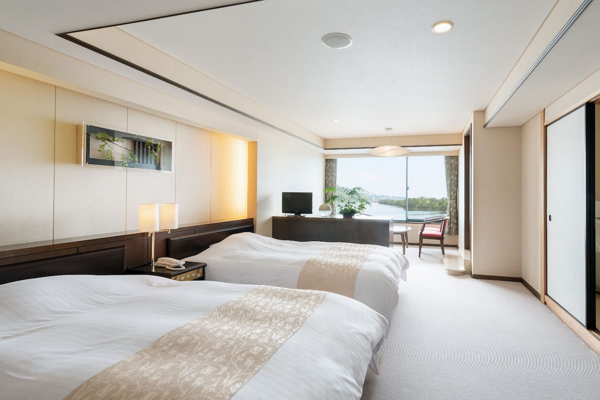 A hotel room with two beds, a desk, a chair, a TV, and a large window offering a view.