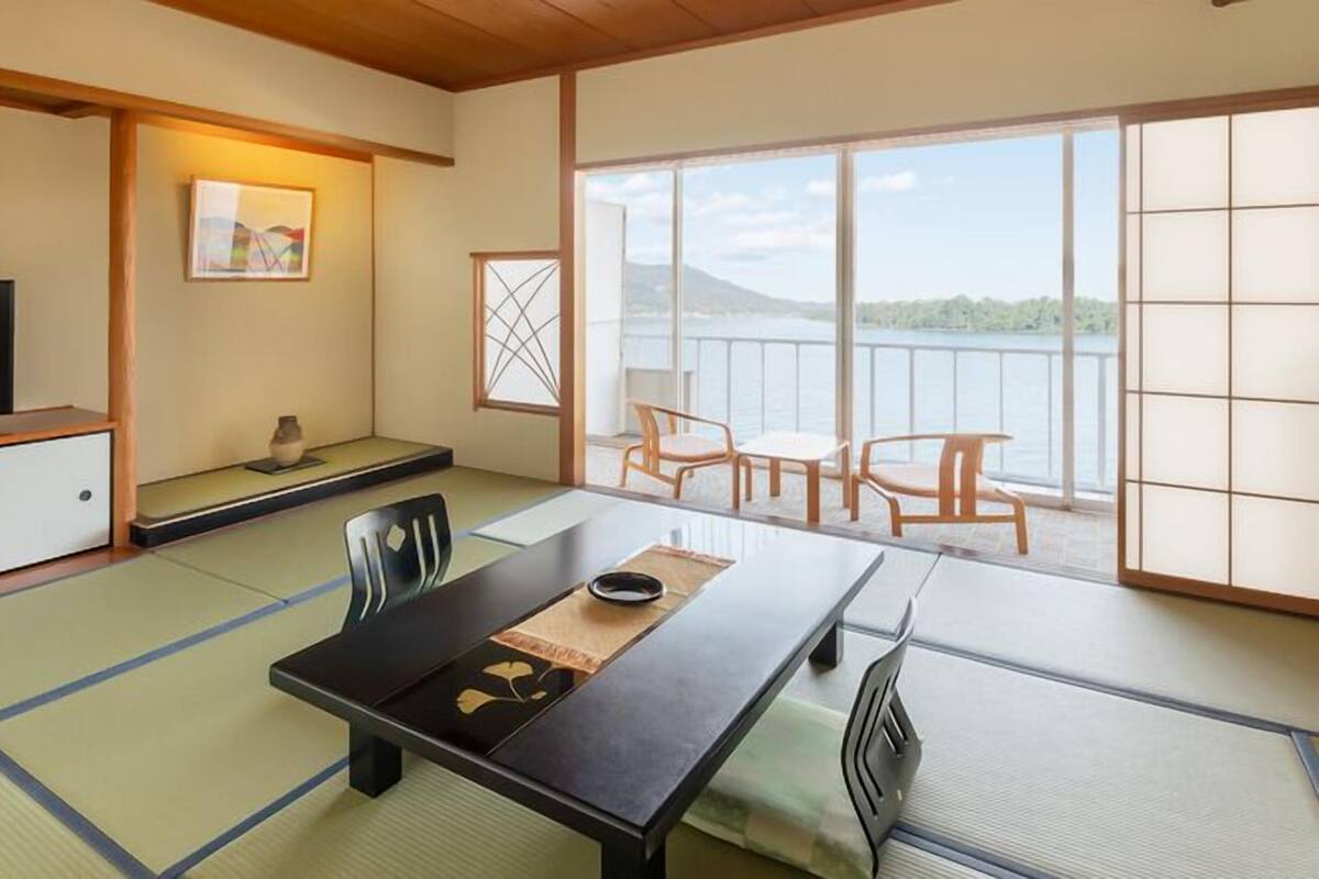 A traditional Japanese room with tatami flooring, a television, a low table with a placemat, and a view of a body of water through large windows.