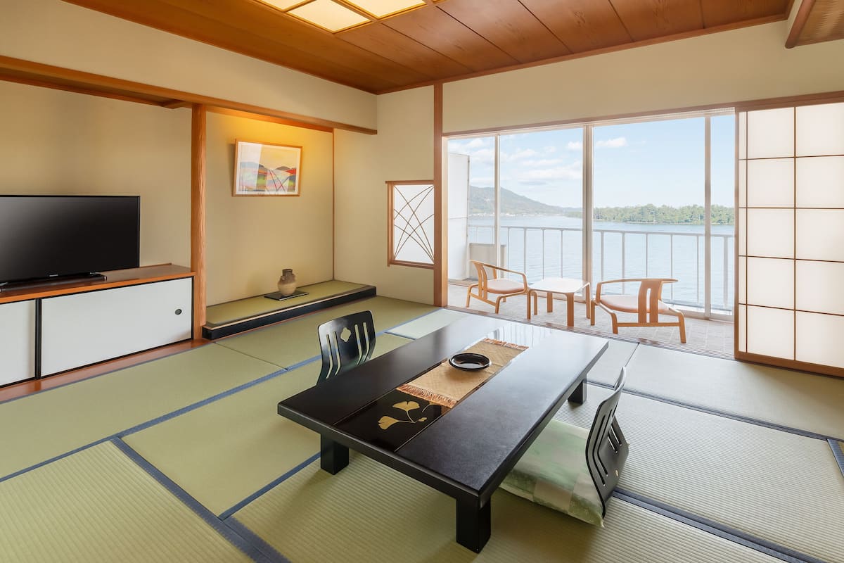 A traditional Japanese room with tatami flooring, a low table with a black mat, a television on a cabinet, and a view of a lake through large windows.