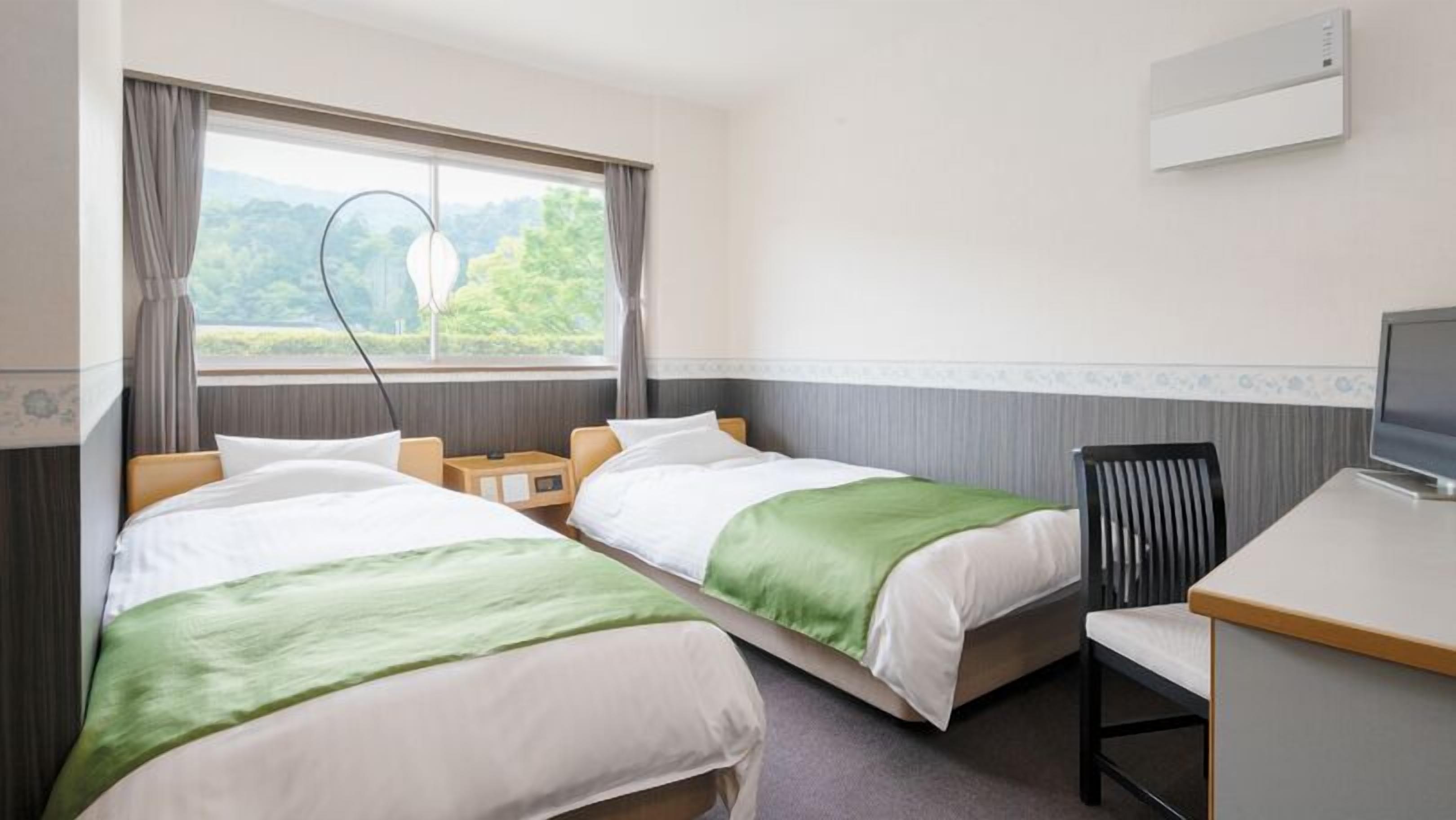 A hotel room with two beds, a desk, a chair, and a window with a view of greenery.