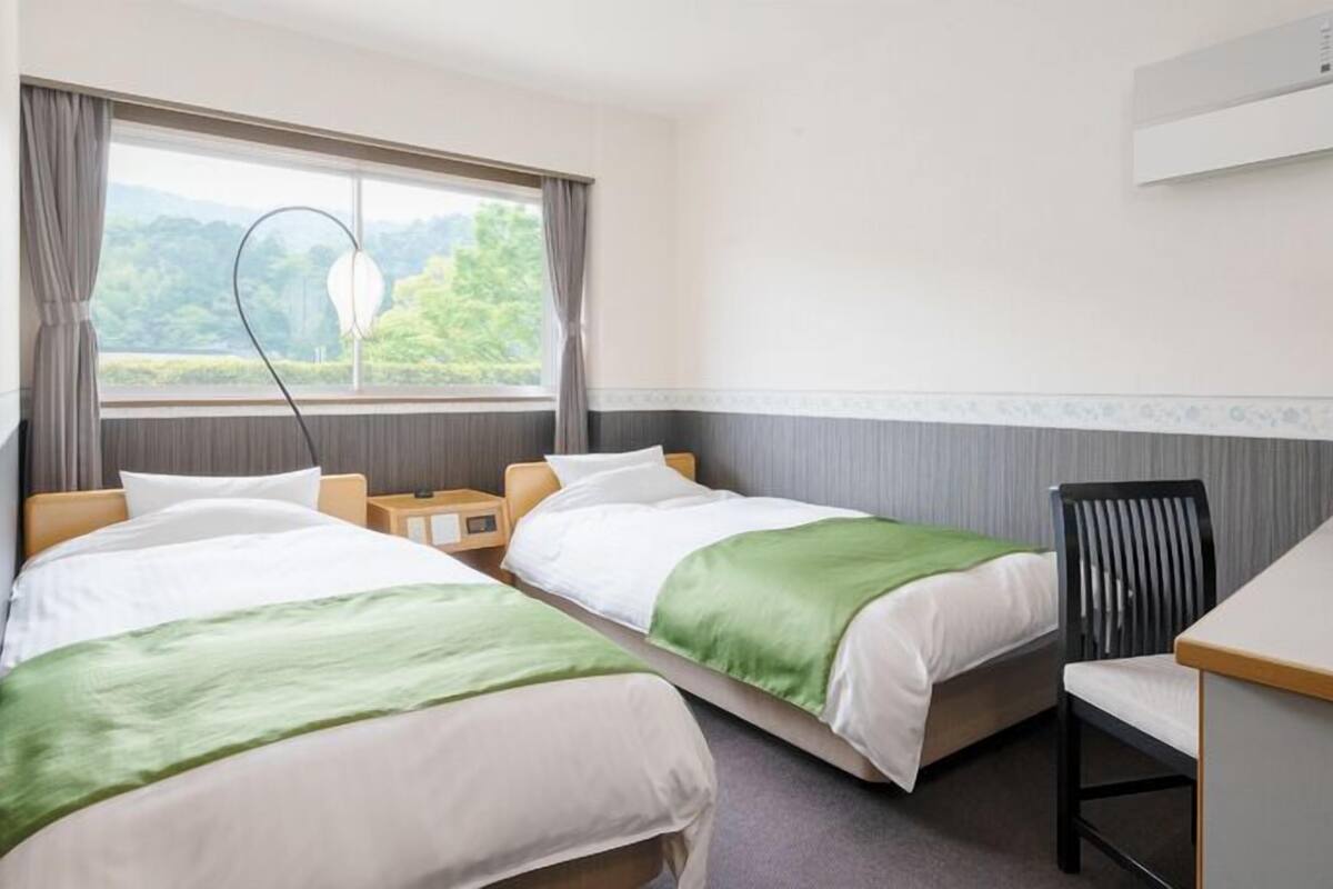 A hotel room with two beds, a desk, a chair, and a window with a view of greenery.