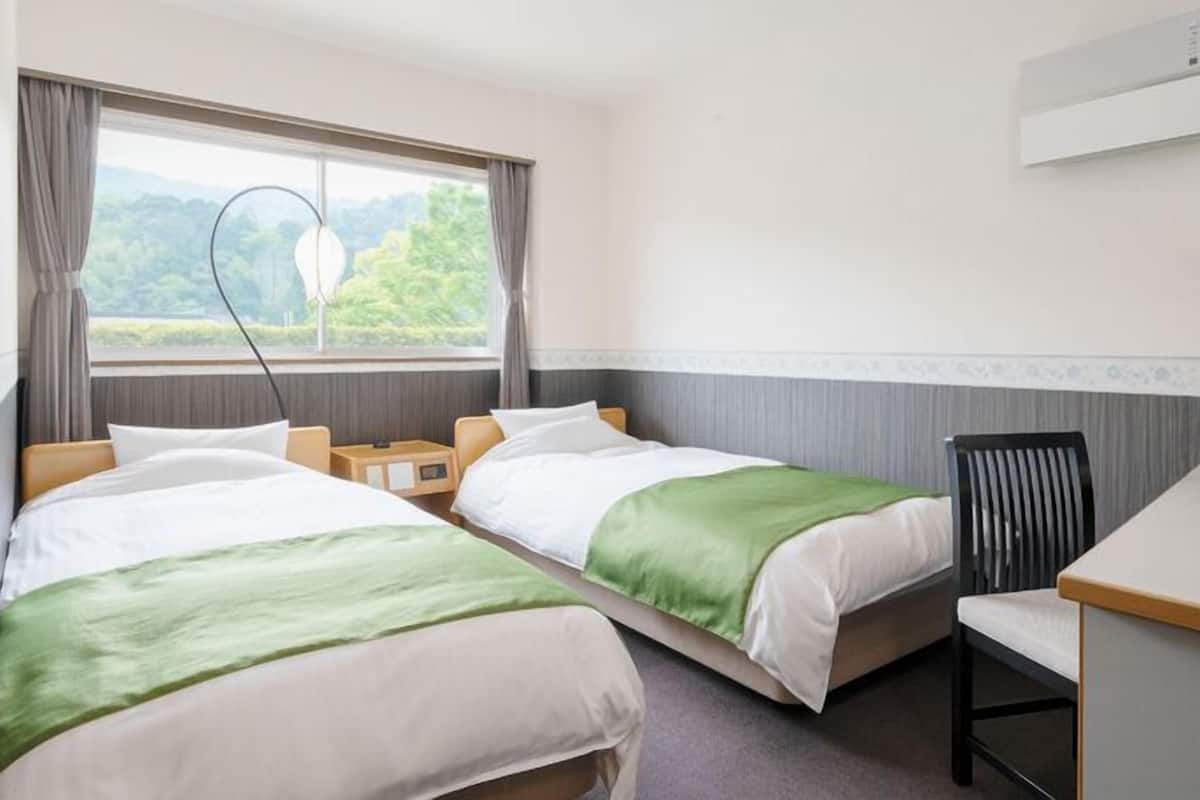 Habitación de hotel con dos camas, un escritorio, una silla y una ventana con vistas a zonas verdes.