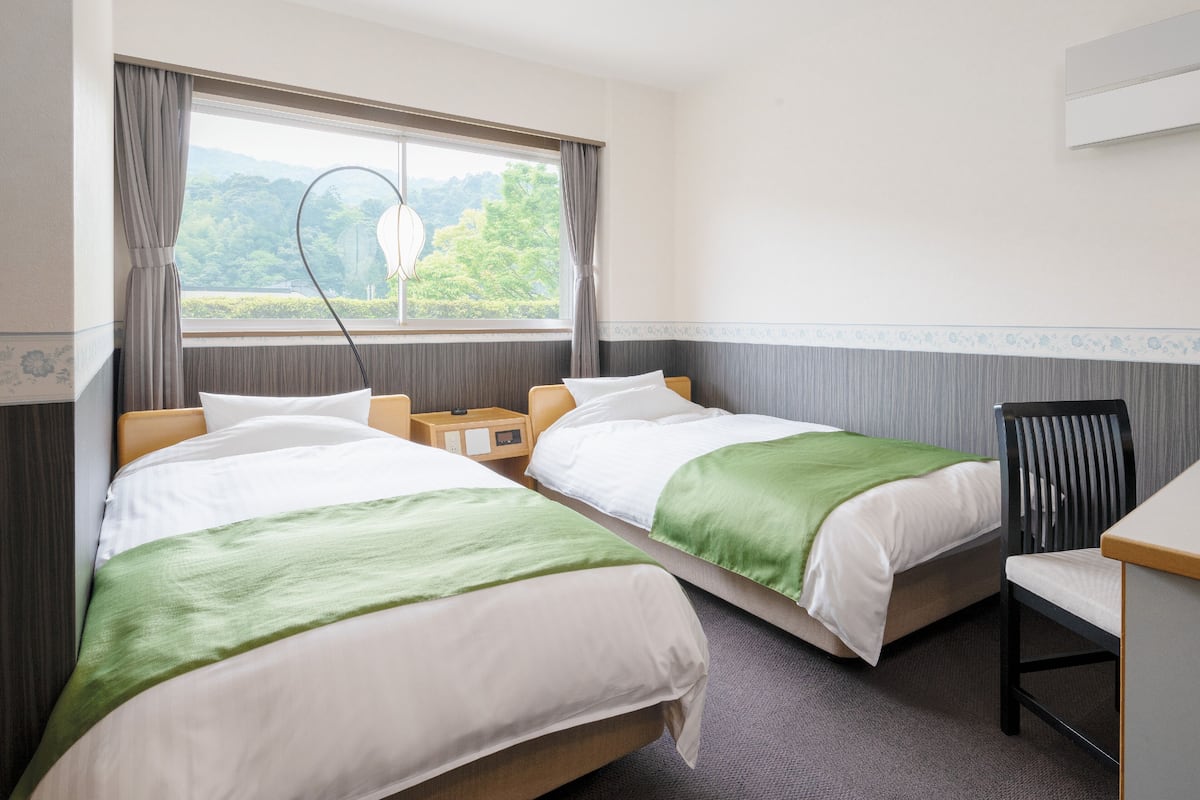 A hotel room with two beds, a desk, and a view of greenery through the window.
