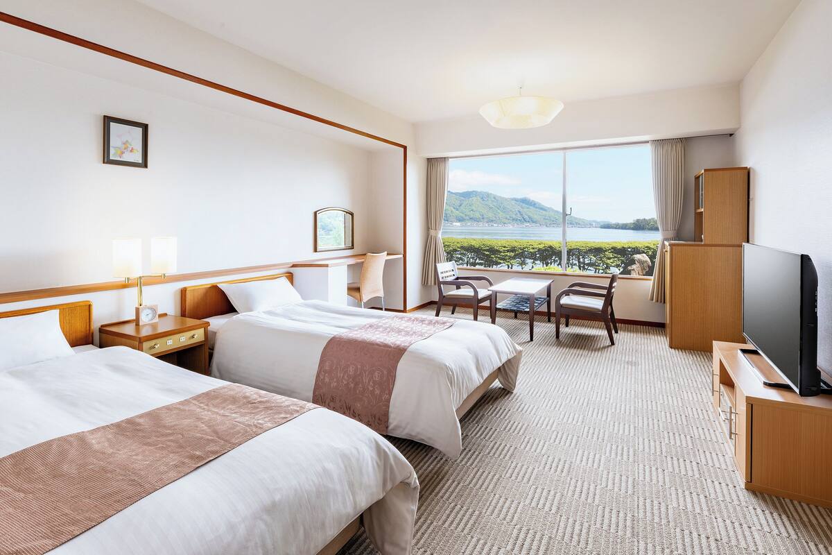 A hotel room with two beds, a TV, a desk, and a view of the outdoors.