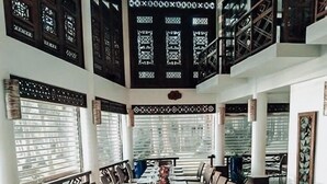 Una larga mesa de comedor dispuesta para una comida, con sillas a ambos lados, grandes ventanales con persianas y un techo alto con elementos decorativos.