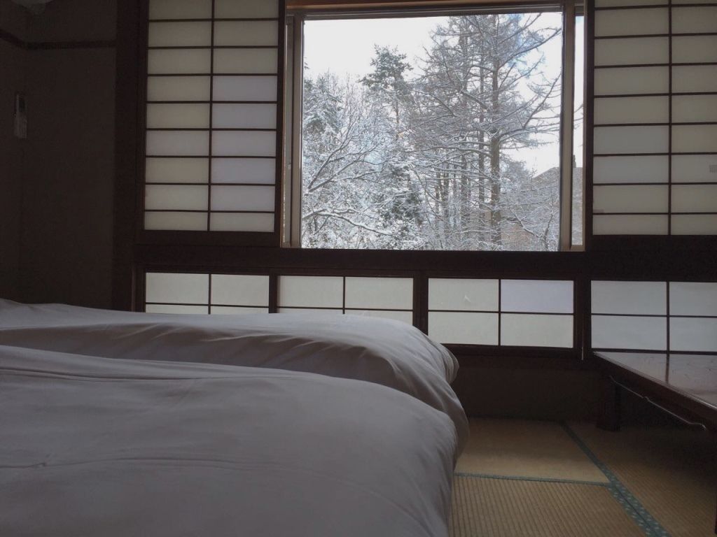 A traditional Japanese room with tatami flooring, a bed, and a window with a view of snow-covered trees.