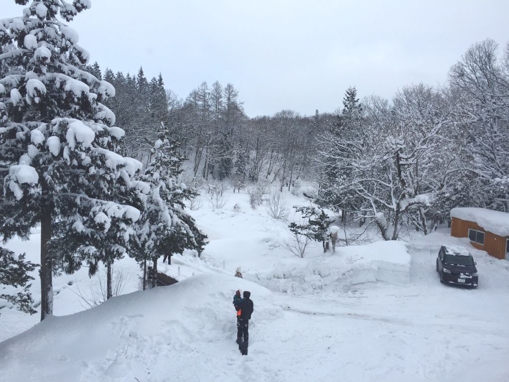 Un paisaje nevado con una persona caminando por un sendero, un coche aparcado a la derecha y un edificio al fondo.