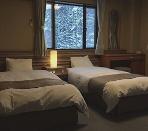 A hotel room with two beds, a nightstand with a lamp, a desk, and a window with a view of snow-covered trees. - Hakuba Landmark Iwatake Lodge (Hakuba)