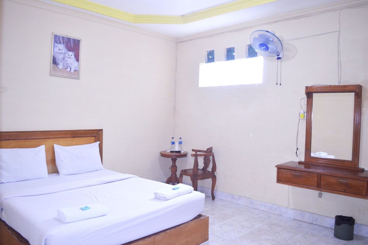 Una habitación de hotel con una cama, un aparador de madera, una silla, una mesita con botellas, un ventilador y una ventana.