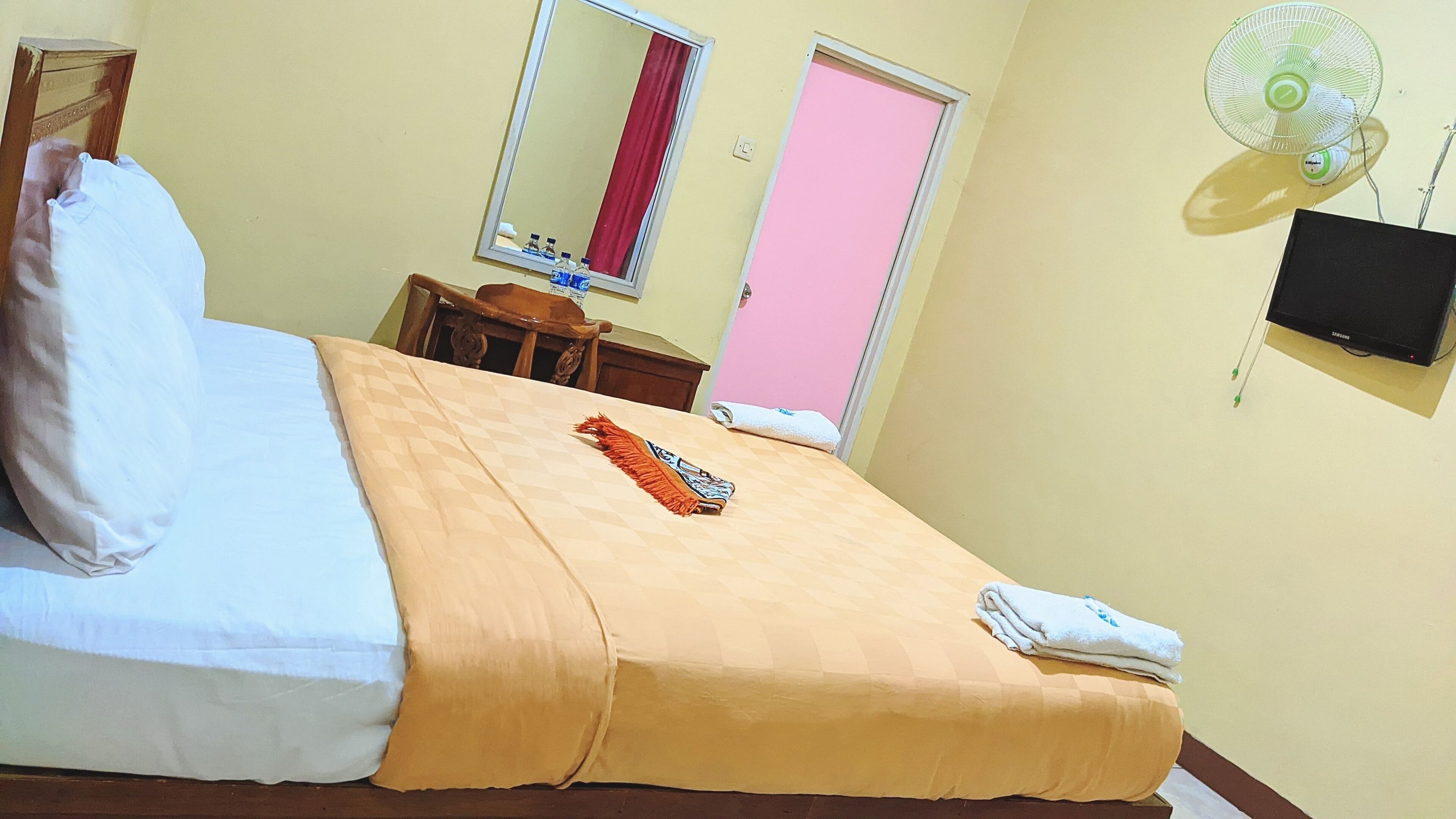 A neatly made bed with a checkered bedspread, a folded towel, and a decorative pillow. A small wooden table with a chair is beside the bed. A mirror hangs on the wall, and a TV is mounted on the wall.