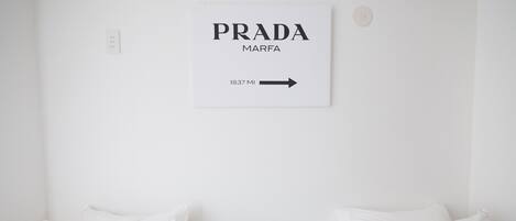 A minimalist hotel room with two beds, a small bedside table with a plant, and a wall sign reading "PRADA MARFA 1837 MI".