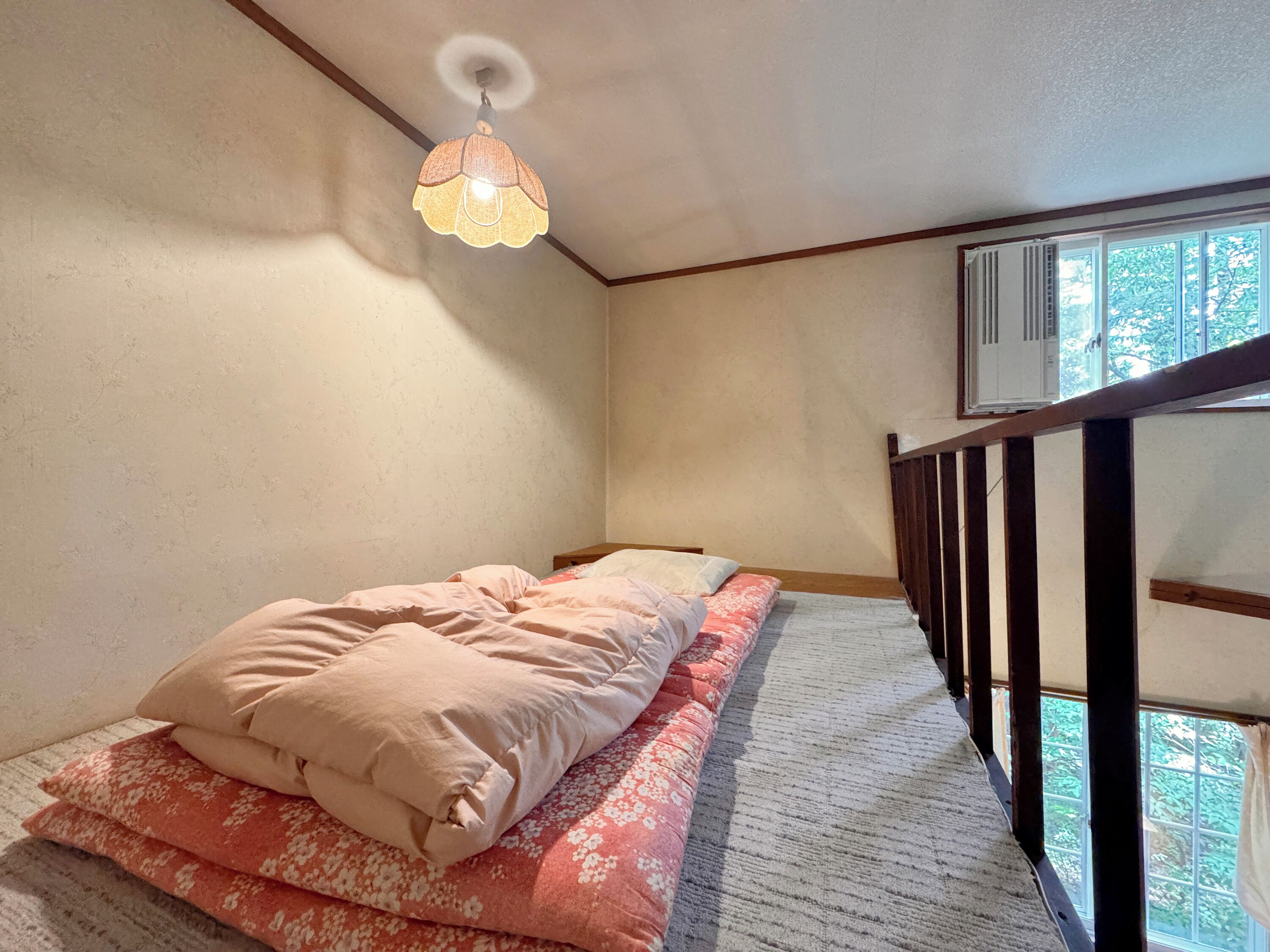 A bedroom with a bed, a hanging lamp, a window with an air conditioner, and a wooden railing.