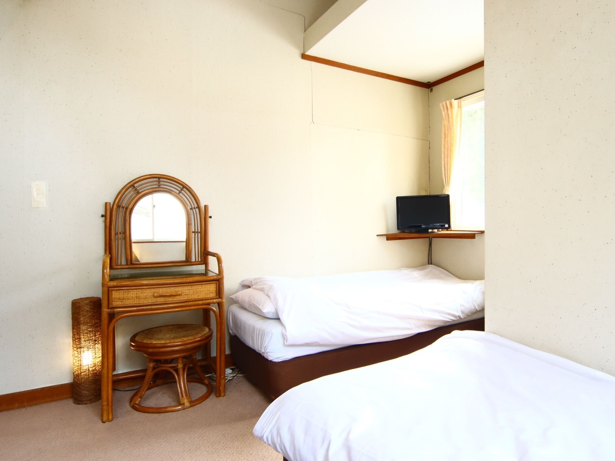 A bedroom with two single beds, a wooden chair with a woven seat, a small table, a mirror, and a TV on a shelf.
