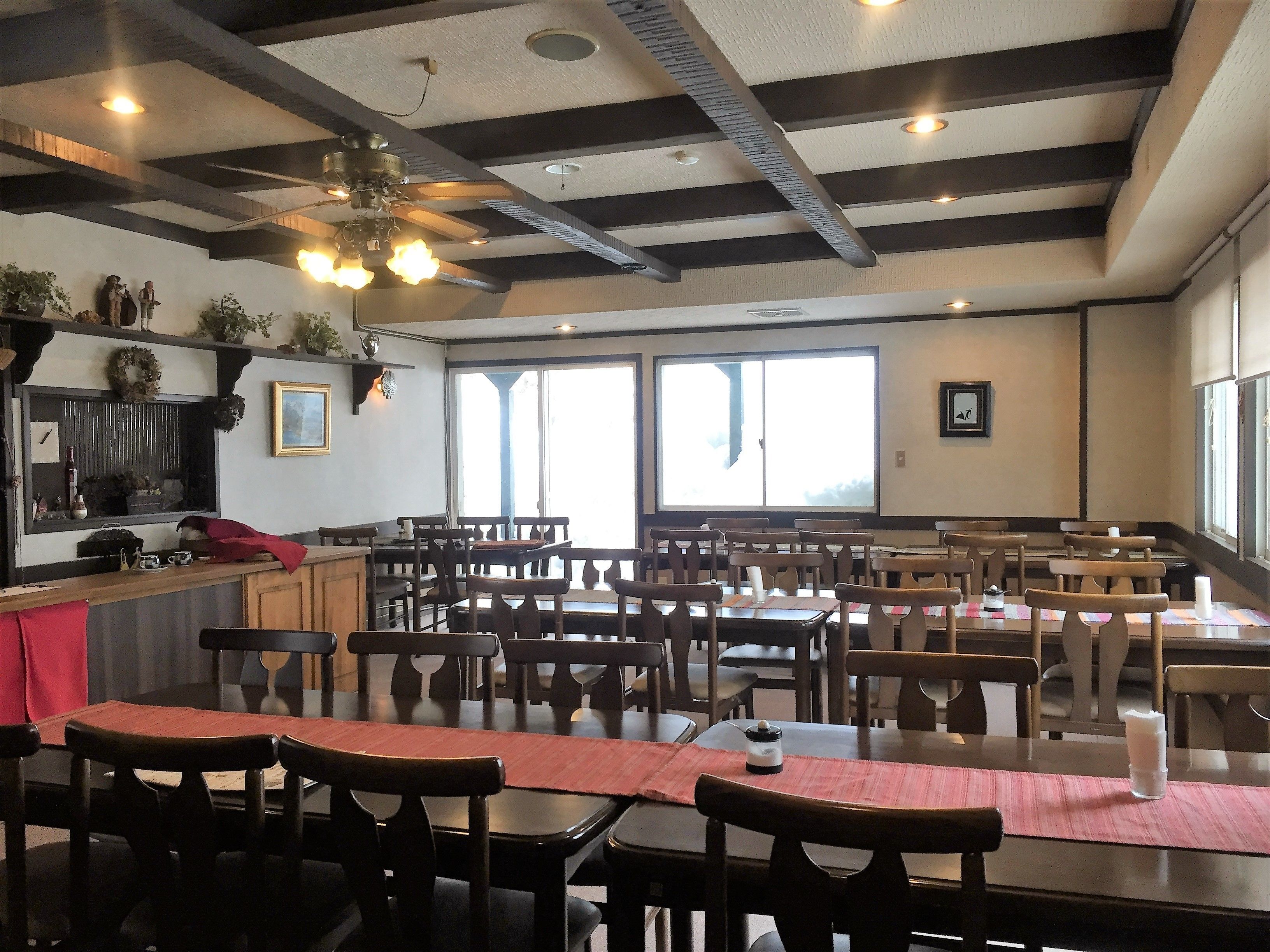 L’intérieur d’un restaurant est aménagé avec des tables et des chaises en bois, un ventilateur de plafond et de grandes fenêtres qui laissent entrer la lumière naturelle.