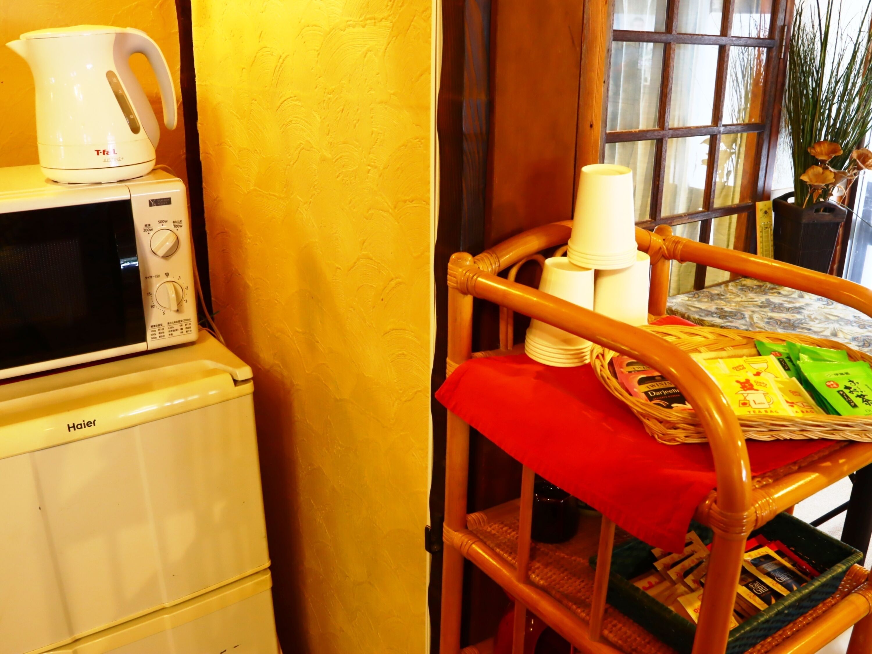A kitchen area with a microwave, kettle, and a wicker chair with a red cloth.