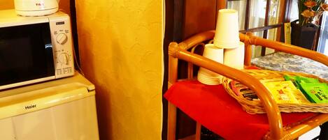 A kitchen area with a microwave, kettle, and a wicker chair with a red cloth.