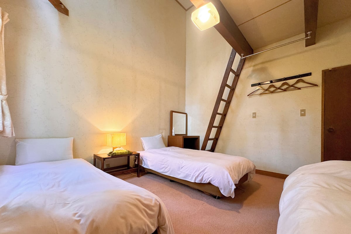 A hotel room with three single beds, a nightstand with a lamp, a wooden ladder, and a mirror on the wall.