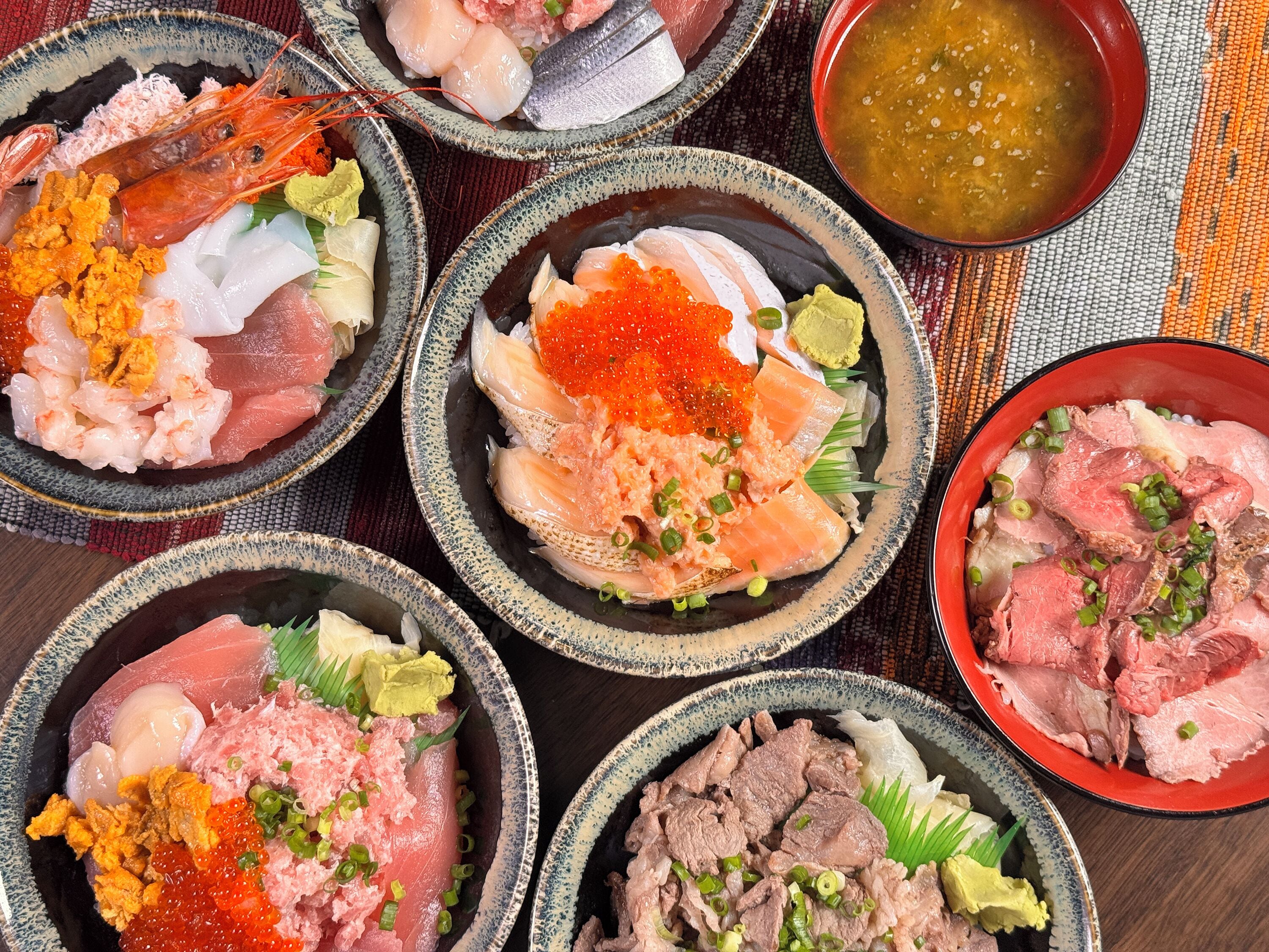 Une variété de plats de fruits de mer servis dans des bols, notamment des sashimis et des sushis, avec un bol de soupe en arrière-plan.
