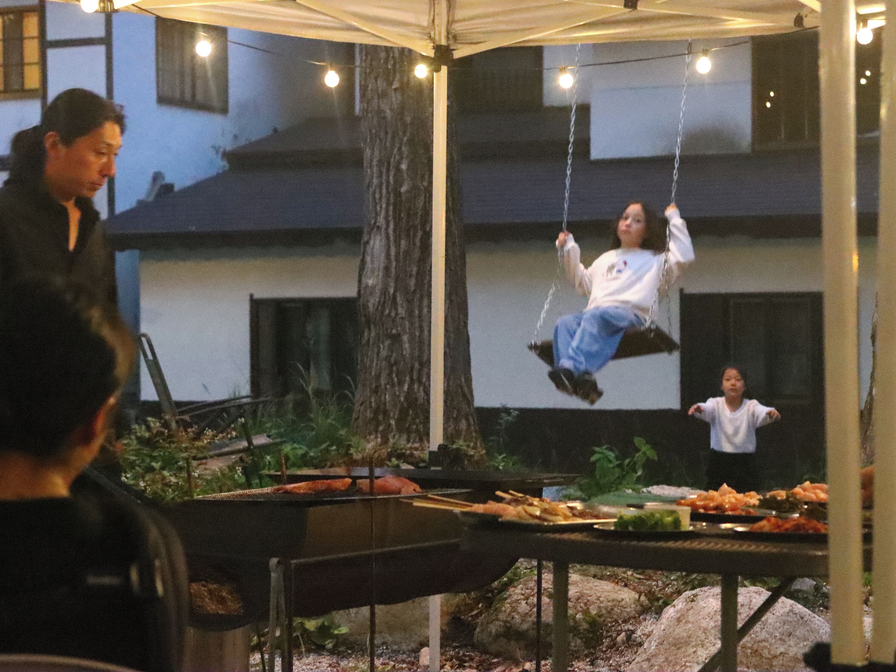 An outdoor dining area with tables and chairs, a swing set, and a building in the background.