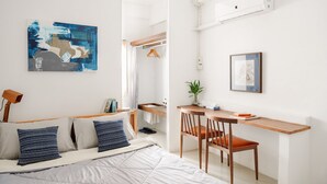Un dormitorio moderno con cama, mesita de noche y escritorio con silla. Hay un espejo, una planta colgante y obras de arte en las paredes.