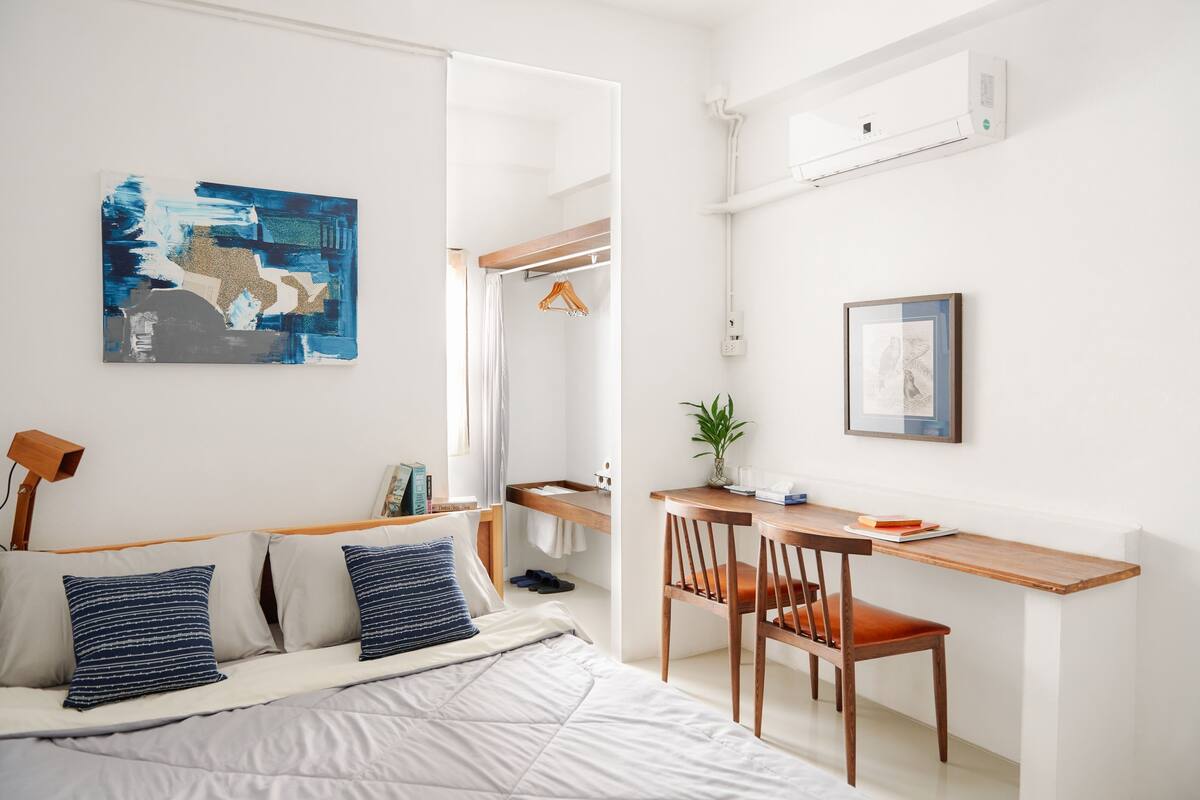 A modern bedroom with a bed, bedside table, and a desk with a chair. There is a mirror, a hanging plant, and artwork on the walls.