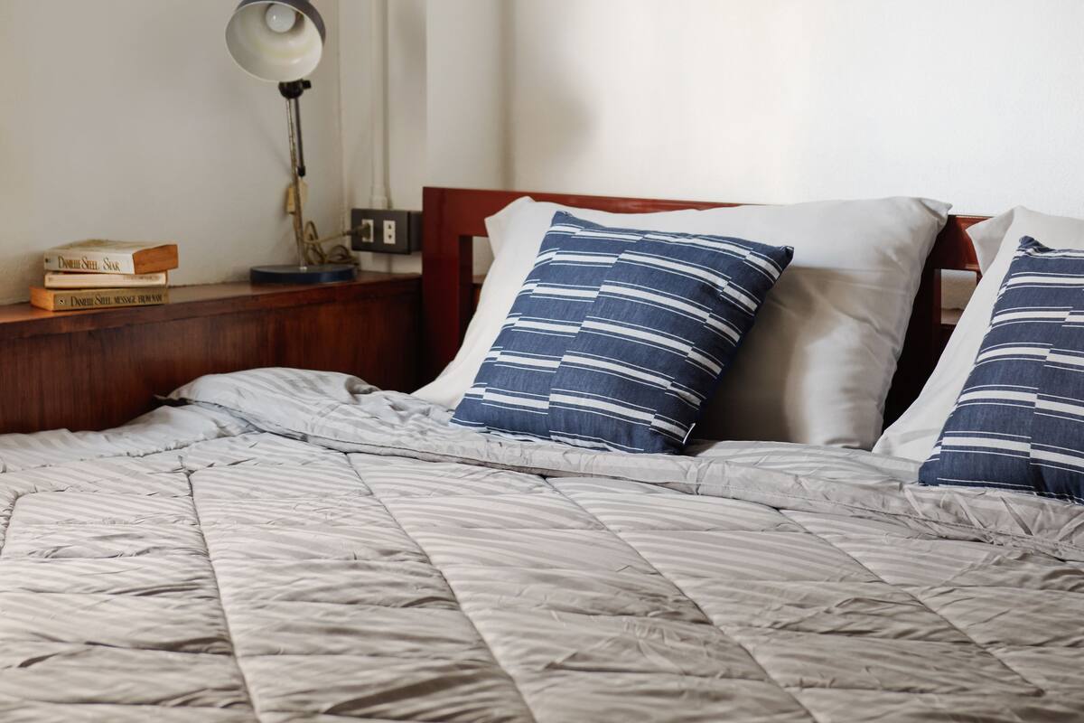 A neatly made bed with a striped comforter, two pillows with blue and white stripes, a bedside lamp, and a stack of books on a wooden nightstand.