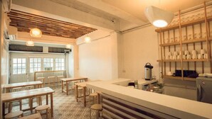 Un café con mesas y sillas de madera, una barra con una máquina de café, y estantes con tazas y platos.