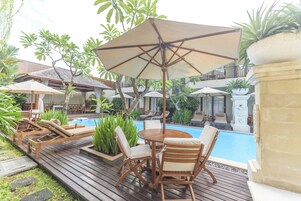 Uma área à beira da piscina com espreguiçadeiras de madeira, uma mesa redonda e guarda-sóis.