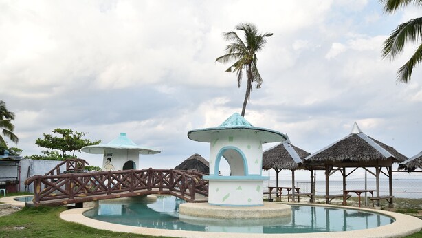 A pool area with a bridge, palm trees, and thatched-roof structures. - King Arthur's Place (Alcoy)