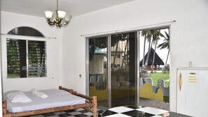 A room with a checkered floor, a bed with white linens, a wooden headboard, a white wardrobe, and a sliding glass door leading to an outdoor area with a thatched-roof structure and palm trees. - King Arthur's Place (Alcoy)