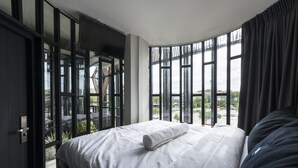 A bedroom with a large bed, a TV, and a view of a cityscape through large windows. - Chingcha Bangkok - Hostel (Bangkok)