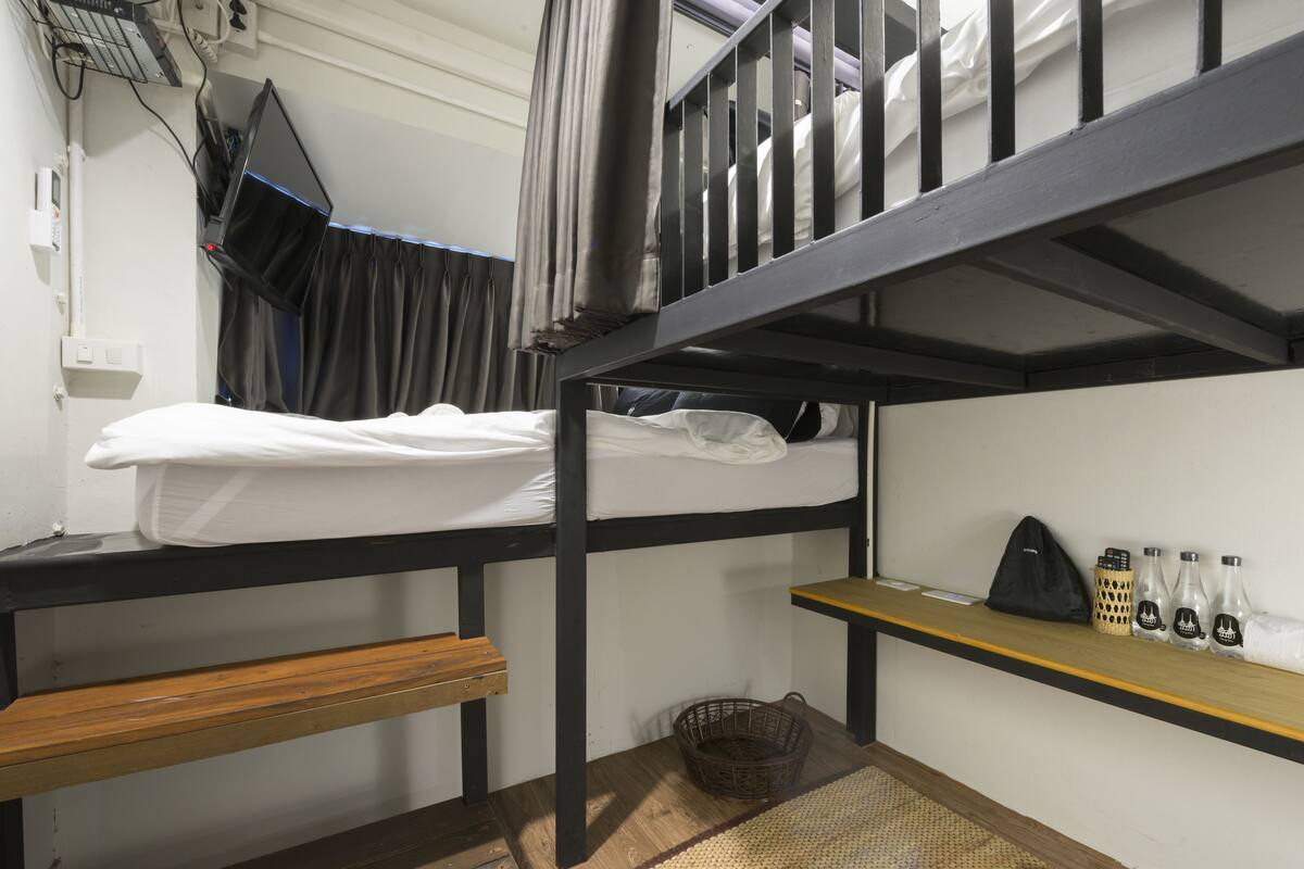Une chambre avec des lits superposés, un banc en bois, une petite étagère avec des bouteilles et un panier posé sur le sol.