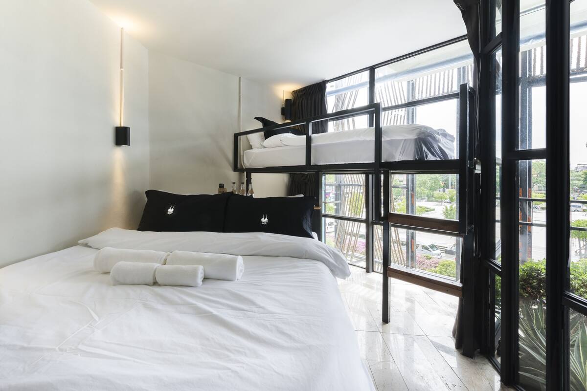 Une chambre d’hôtel moderne avec un lit superposé, une grande fenêtre avec des rideaux et une table de chevet avec une lampe.